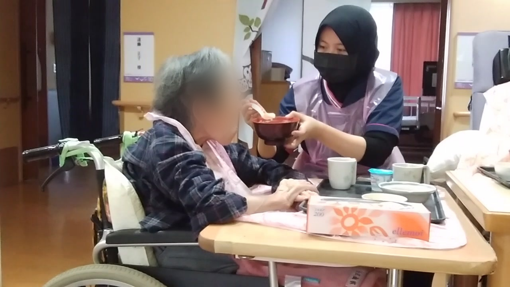 介護施設で、インドネシア人の特定技能介護職員が車いすの利用者さんの食事介助をしている様子。お椀を手に持ち、スプーンで汁物を口元へ運んでいる。