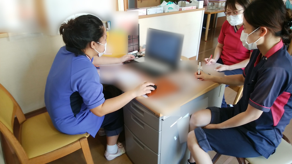 介護施設の職員スペースで、インドネシア人介護職員がノートパソコンを操作しながら日本人の介護職員2人と打ち合わせをしている様子。日本人職員はメモを取りながら話を聞いている。